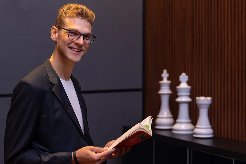 Ricardo Werlang segurando um livro ao lado de peças de xadrez brancas.