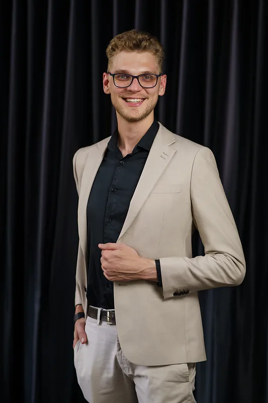 Ricardo Werlang sorrindo, blazer bege e camisa preta, cortina escura ao fundo.