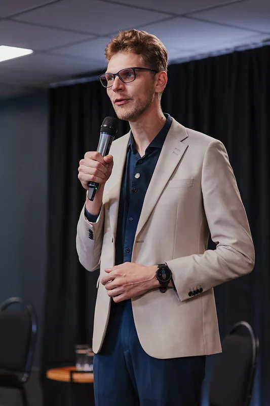 Ricardo Werlang falando ao microfone, blazer bege, fundo escuro.