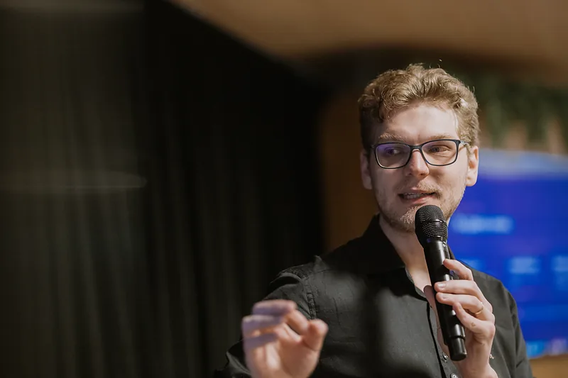 Ricardo Werlang em primeiro plano com microfone, fundo desfocado.